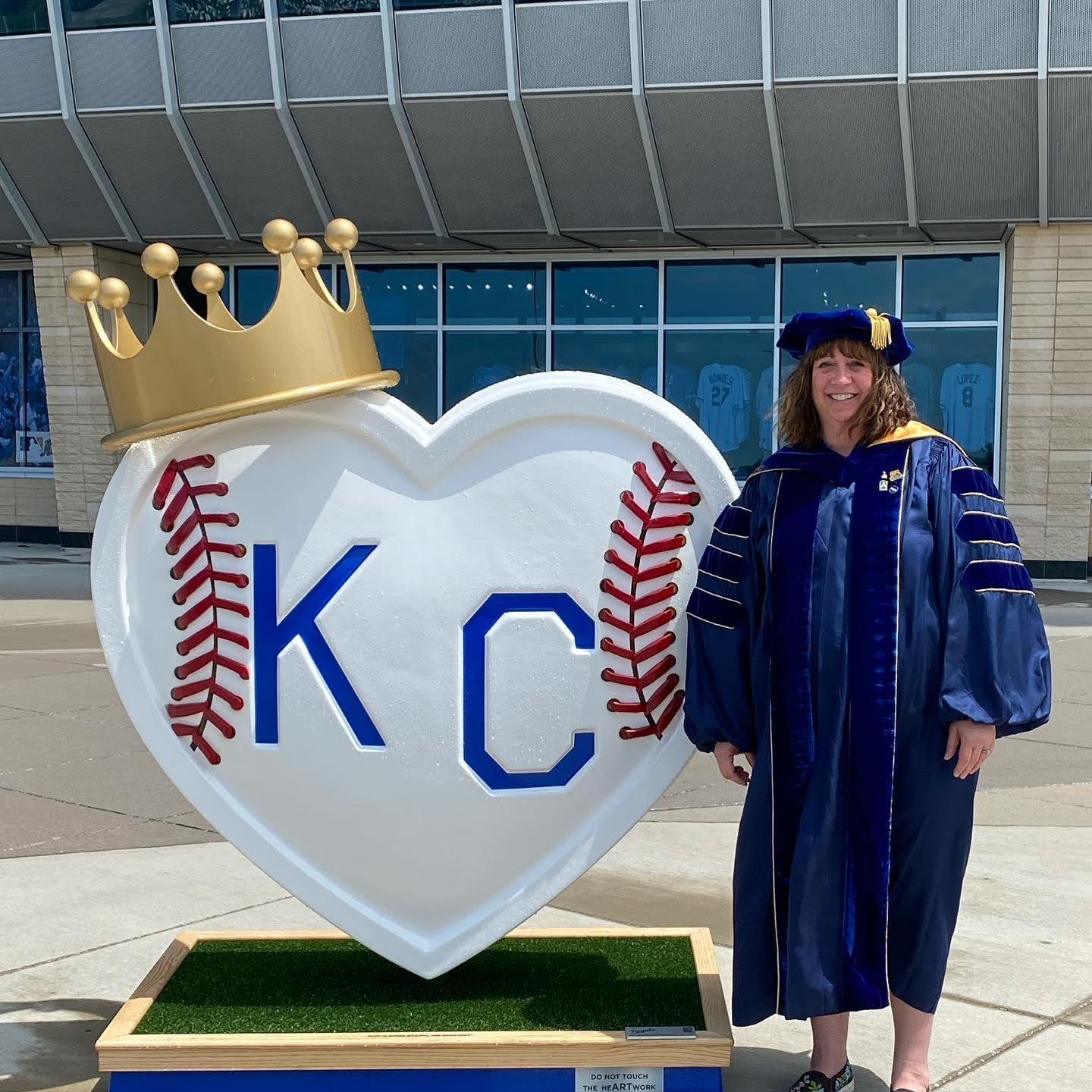 Tiffani Riggers-Piehl standing in doctoral regalia next to a KC heart painted to look like a baseball