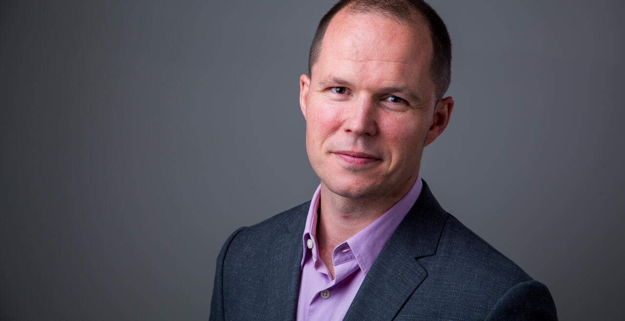A head shot of Matthew Edwards, a white man with short brown hair in a grew stuit jacket and a light blue shirt. 