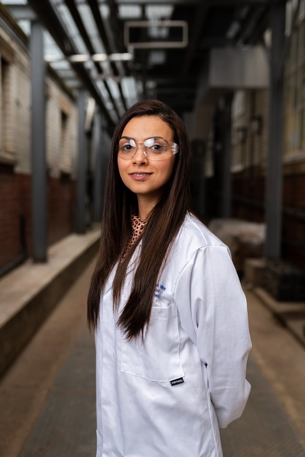 Person looking directly at camera with long brown hair, wearing goggles and white lab coat